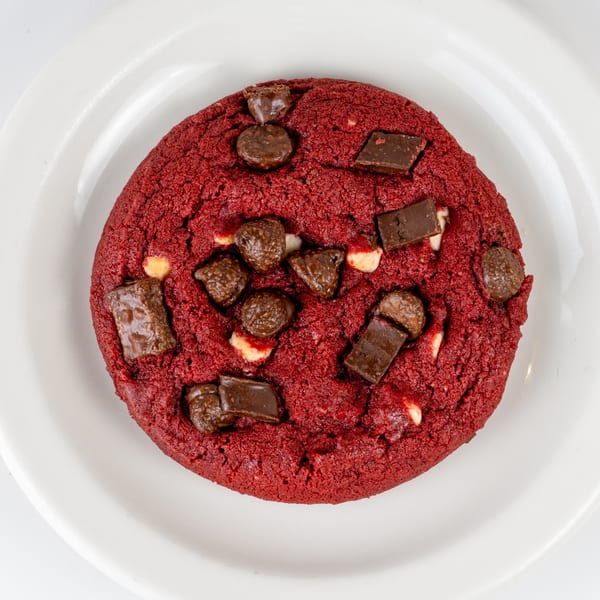 A vibrant red velvet cookie with chocolate chunks and chips on a white plate.