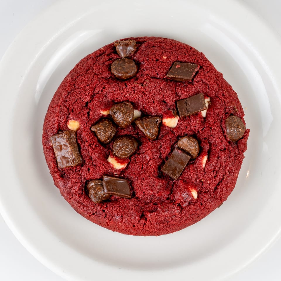 A vibrant red velvet cookie with chocolate chunks and chips on a white plate.