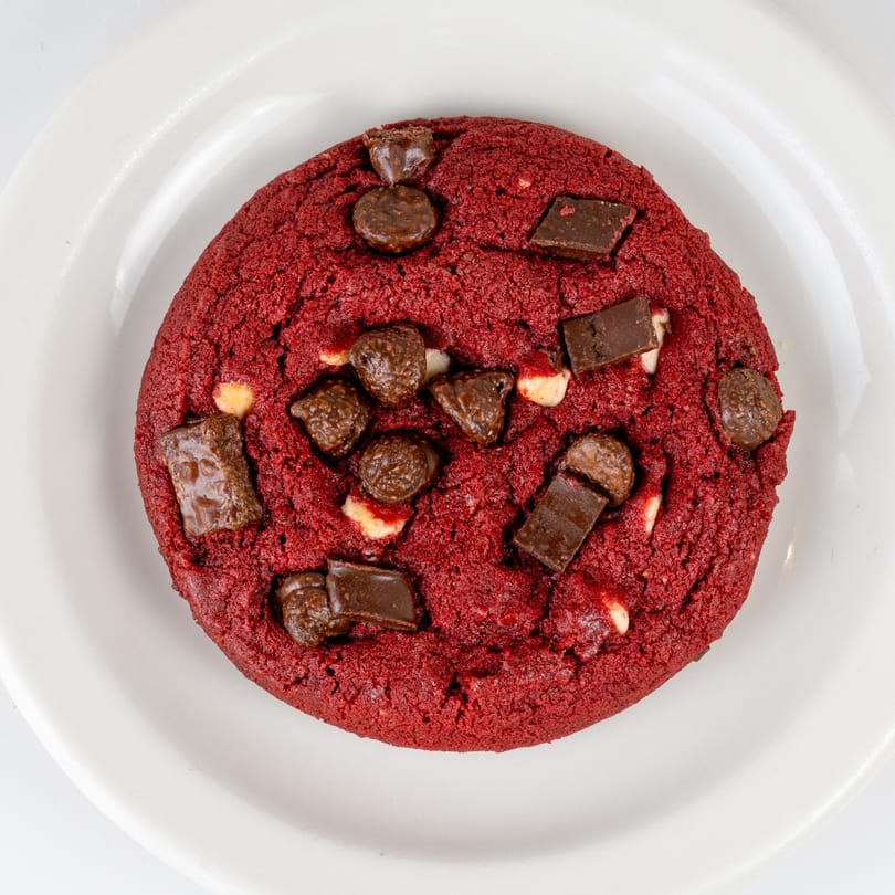 A vibrant red velvet cookie with chocolate chunks and chips on a white plate.
