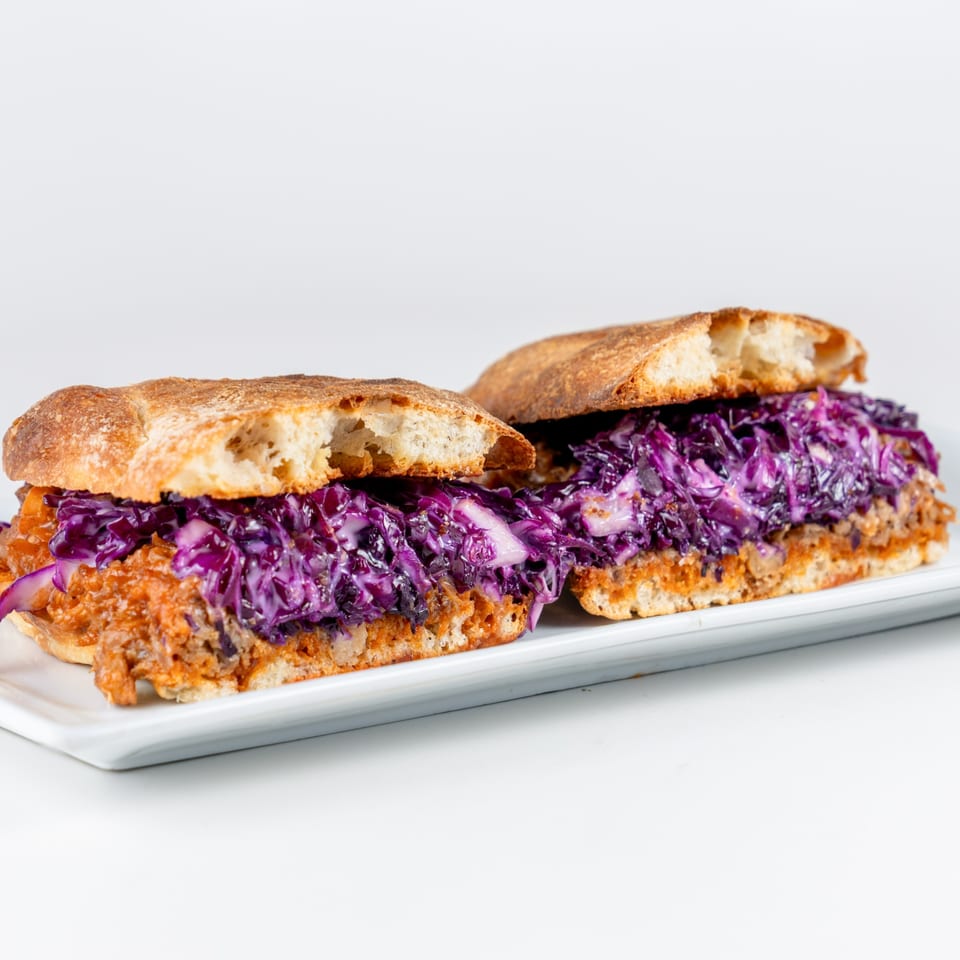 Two pulled pork sandwiches with purple cabbage slaw on ciabatta bread, served on a white platter.
