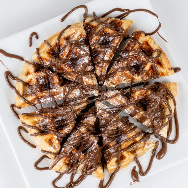 Round waffle cut into slices, drizzled with chocolate sauce and powdered sugar on a white plate.