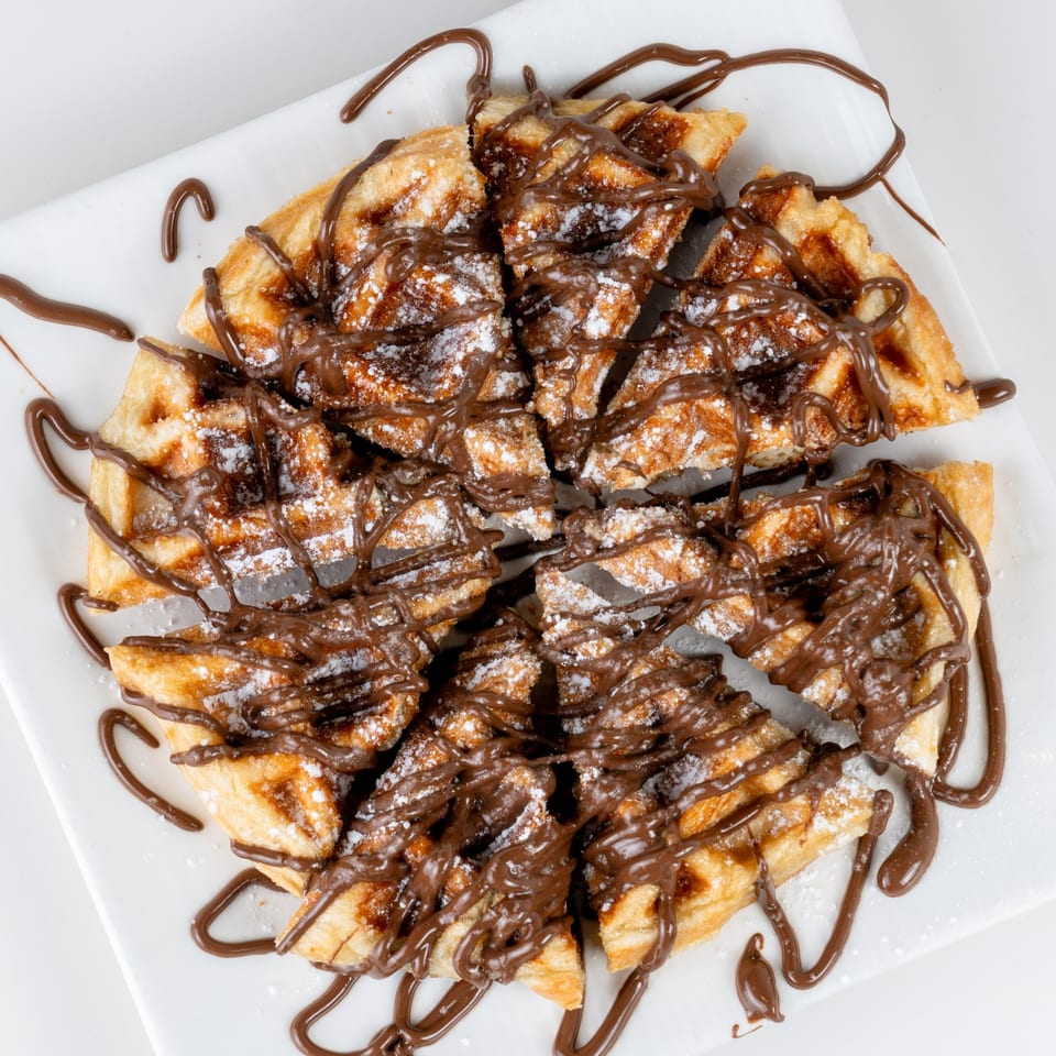 Round waffle cut into slices, drizzled with chocolate sauce and powdered sugar on a white plate.