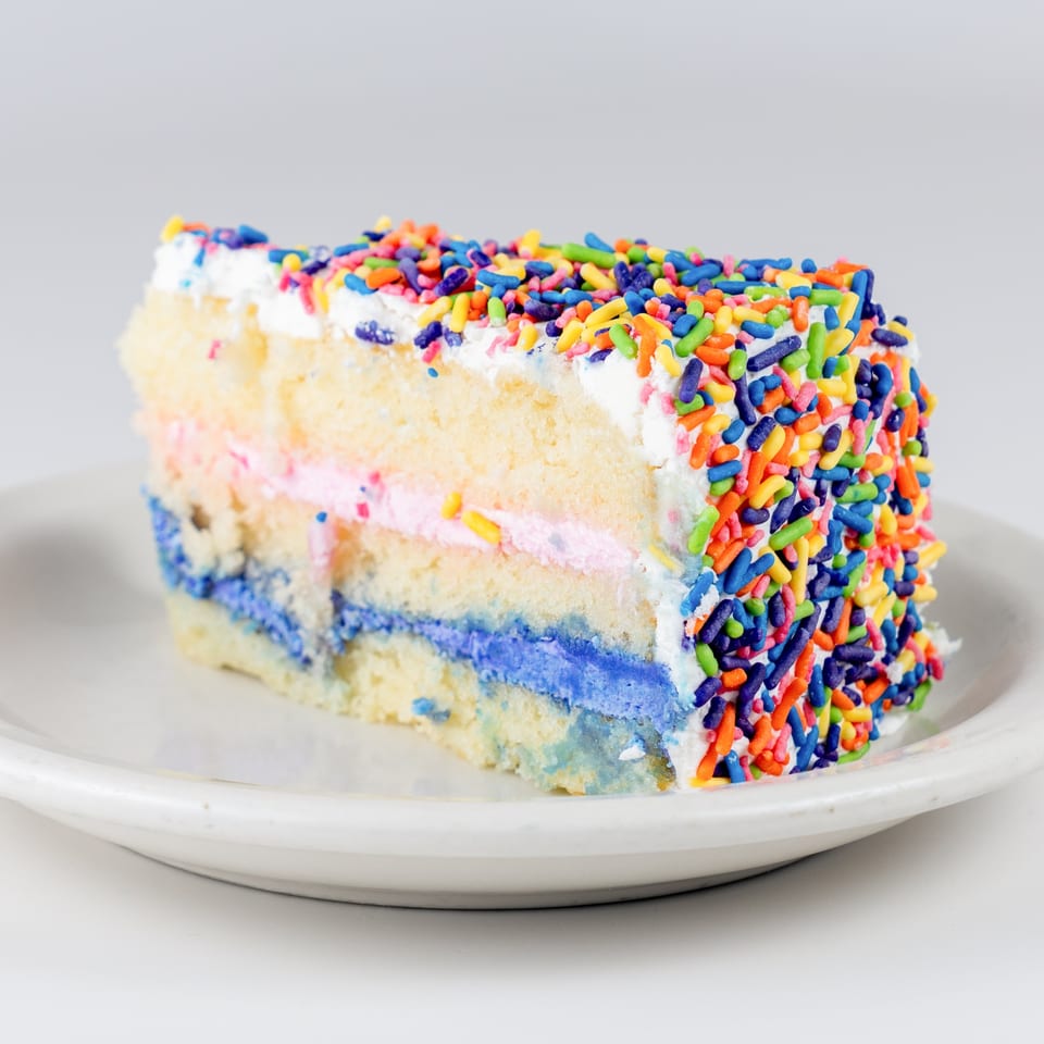 Slice of Kudo Society Cafe's vanilla cake with colorful sprinkles, pink and blue frosting layers.