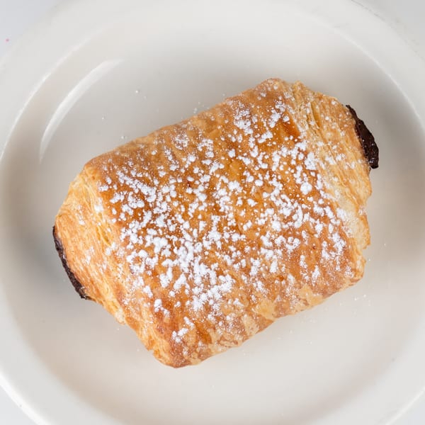 A golden-brown pain au chocolat, dusted with powdered sugar, rests on a white plate.