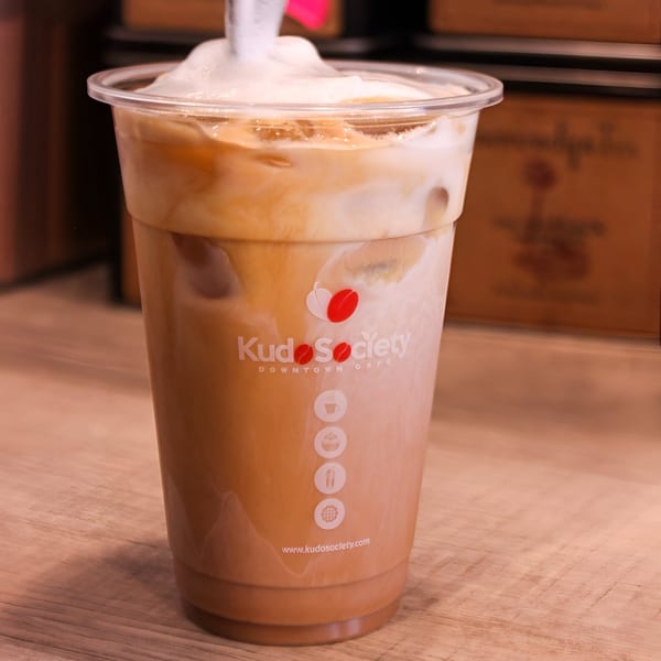 Iced coffee with foam in a Kudo Society Cafe branded cup on a wooden surface.